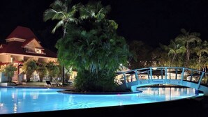 A nighttime view of a resort with a swimming pool, palm trees, and a bridge.
