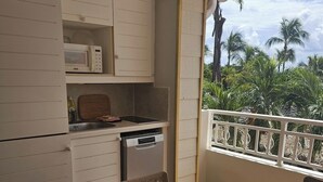A compact kitchen with a microwave, stove, and sink, a dining table with chairs, and a balcony with a railing and palm trees.