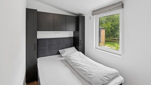 Ett kompakt sovrum med en enkelsäng, inbyggda mörka skåp, ett fönster med utsikt över grönområden och en taklampa.
