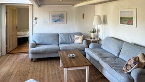 Una sala de estar con un sofá gris, una mesa de centro de madera y una mesita auxiliar con una lámpara. Hay una puerta que da a otra habitación y un cuadro en la pared.