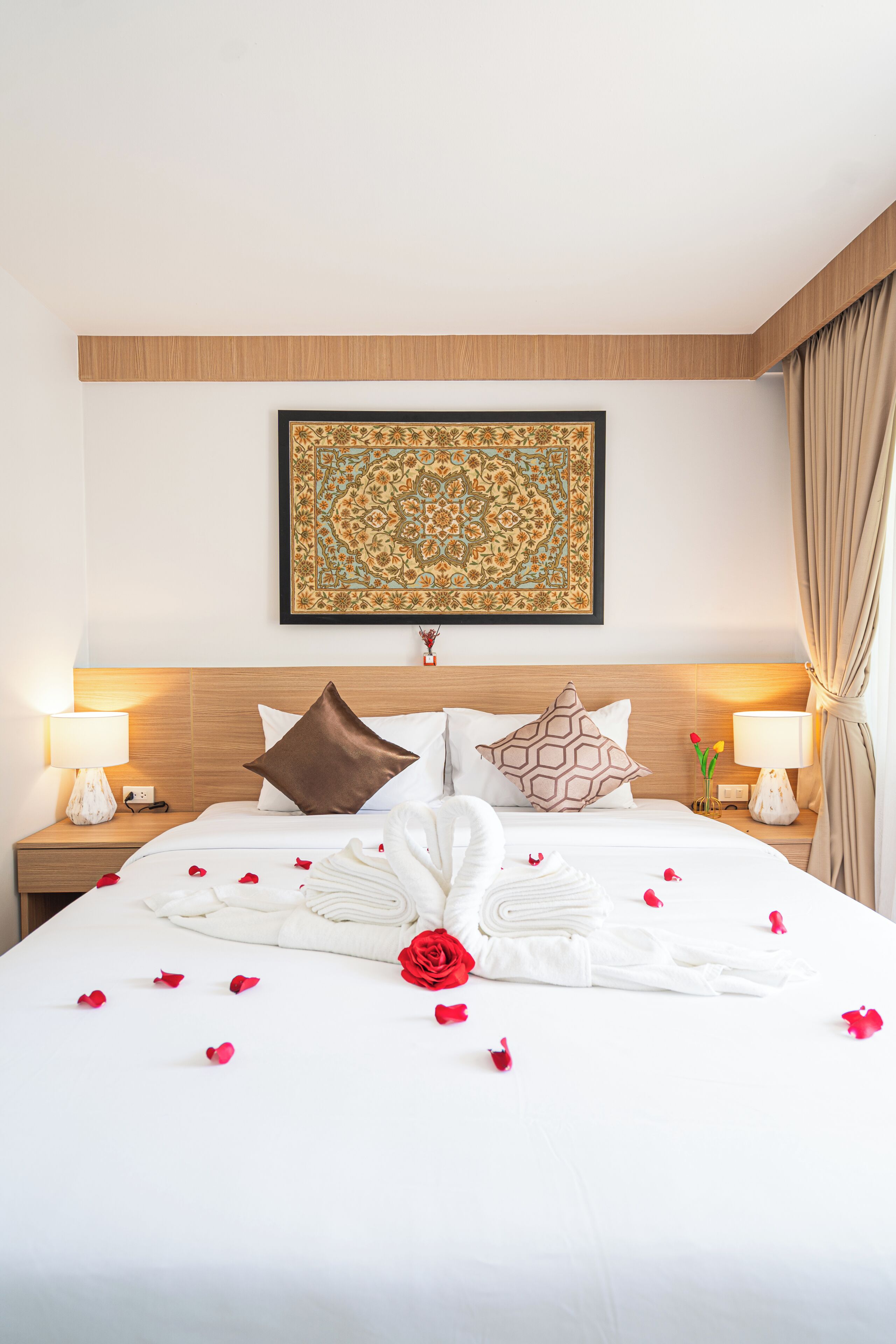 Una cama bien tendida con pétalos de rosa, una obra enmarcada encima y dos lámparas de noche.