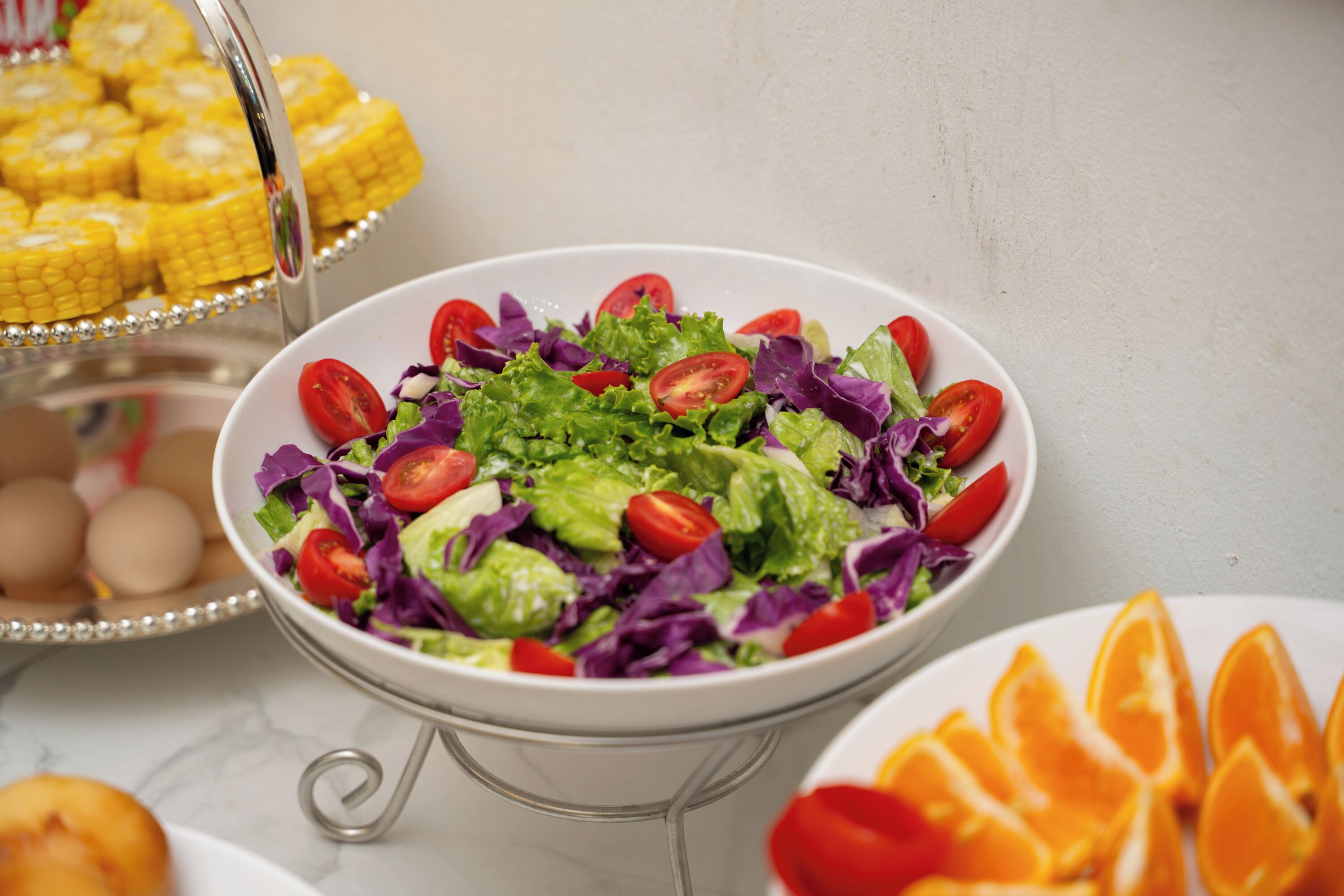 A buffet table with a variety of food items including a salad, corn on the cob, eggs, and orange slices.