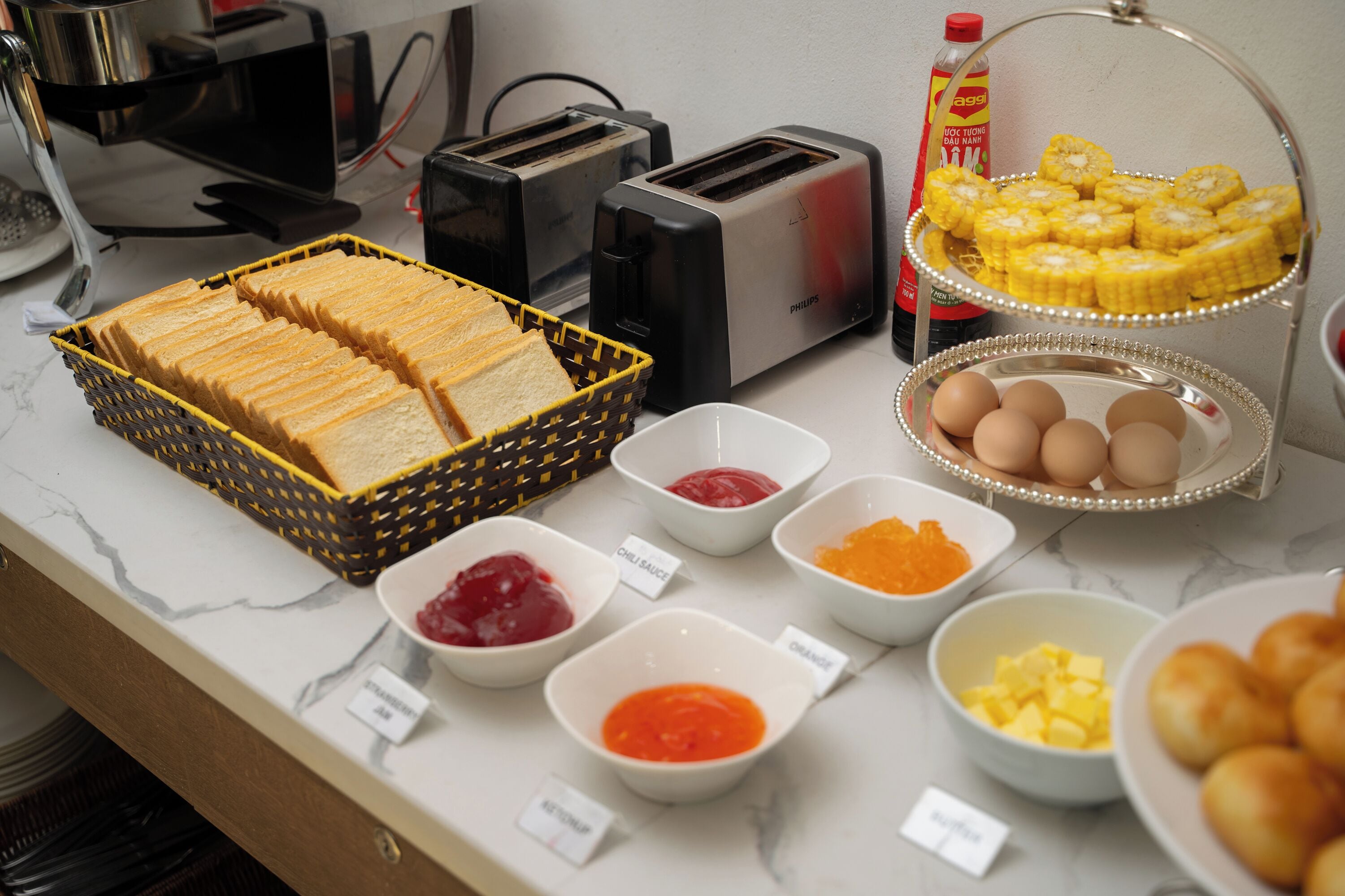 A breakfast buffet with bread, eggs, corn on the cob, and various condiments.