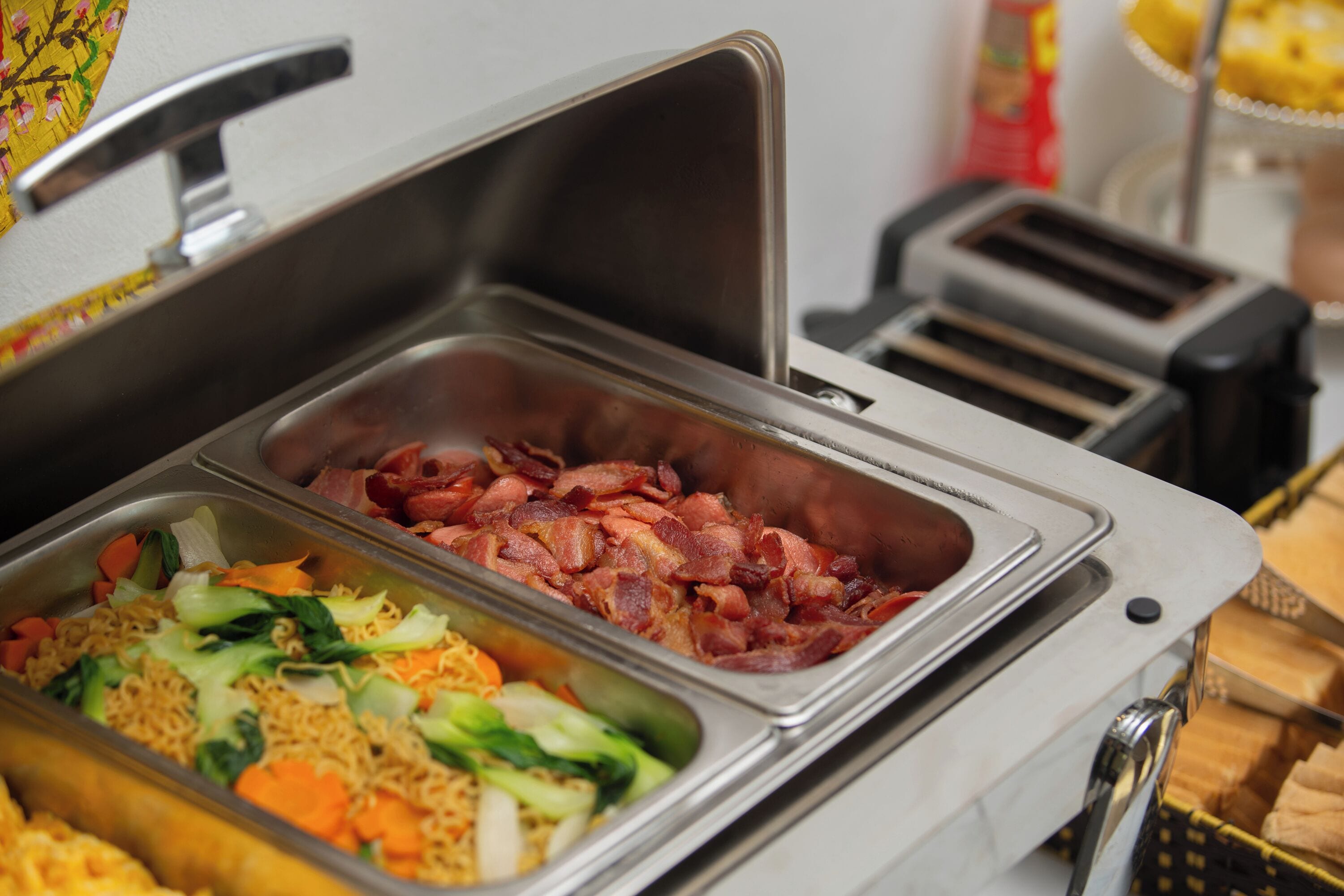 A buffet with various food items including noodles, vegetables, and bacon.