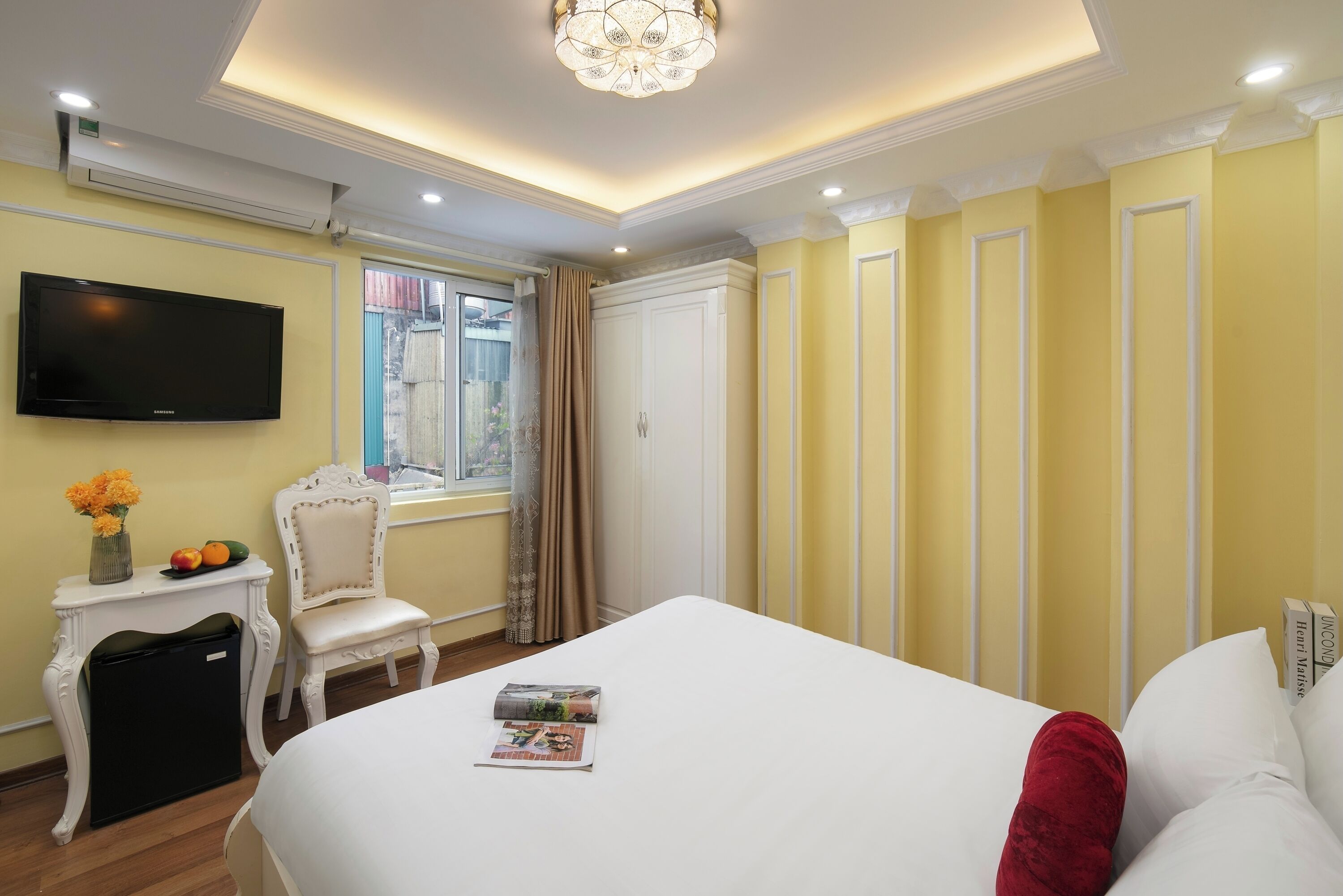 A hotel room with a bed, a TV, a small table with a vase of flowers, a chair, and a window with curtains.