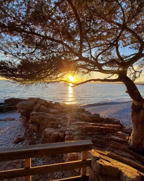 A sunset over a rocky shoreline with a wooden bench in the foreground. - Le Sémaphore (Saint-Raphaël)