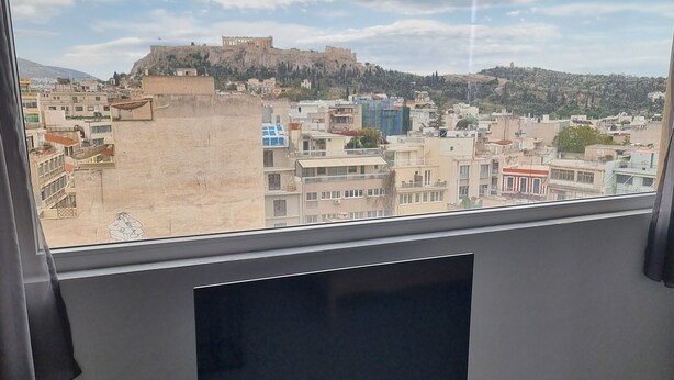 A room with a large window overlooking a cityscape, including a hill with a fort-like structure. - athens parthenon view (Athens)