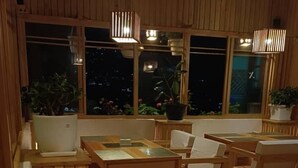 A cozy, wooden-furnished dining area with tables and benches, large windows with potted plants, and hanging pendant lights. - UV INN (Thimphu)