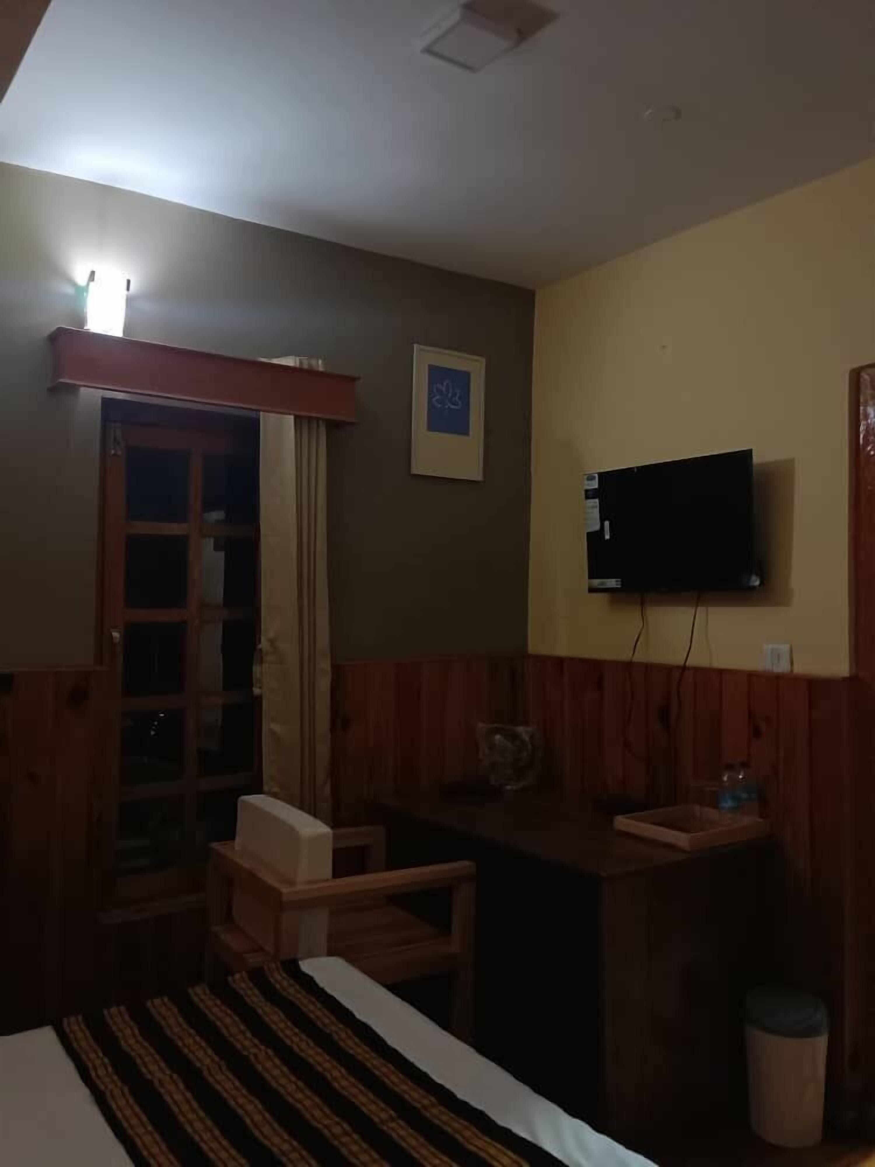 A small, dimly lit room with a bed, a wooden desk, a television mounted on the wall, and a window with curtains.