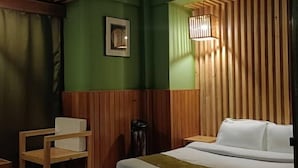 A hotel room with a bed, a chair, a small table, and a wall-mounted light fixture. - UV INN (Thimphu)