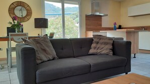 Un salon moderne avec un canapé gris, une petite table, un téléviseur, un coin cuisine doté de meubles en bois et une grande fenêtre offrant une vue sur la verdure et les collines.
