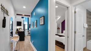 A hotel room with a blue accent wall, a bed, a bathroom with a glass shower, and a hallway with artwork and a desk.