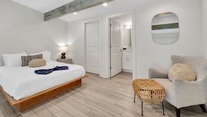 Una camera da letto con un letto in legno, una poltrona grigia, un poggiapiedi in vimini e uno specchio rotondo appeso al muro.