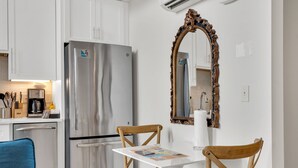 A compact kitchen with white cabinets, a stainless steel refrigerator, a wooden dining table with two chairs, and a decorative mirror. - Frenchmen St Gem • Walk to French Quarter (New Orleans)
