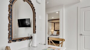 A hotel room with a large ornate mirror, a flat-screen TV, a white table with a magazine, and a wooden chair. - Frenchmen St Gem • Walk to French Quarter (New Orleans)