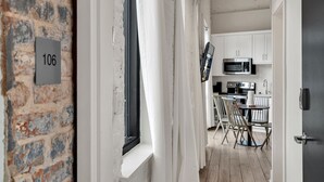 Un couloir étroit, avec un mur de briques sur la gauche et une porte sur la droite. On aperçoit une fenêtre avec des rideaux blancs, et à l’arrière-plan, on distingue un coin cuisine avec des meubles blancs et un four à micro-ondes.