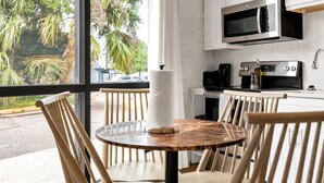 Une cuisine avec des meubles blancs, un four à micro-ondes en acier inoxydable et une machine à café. On y trouve une table ronde avec des chaises, ainsi qu’une fenêtre donnant sur des palmiers et une rue.
