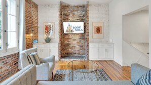 Una sala de estar moderna con chimenea de ladrillo, muebles blancos y un televisor mostrando 'BOOK NOLA'.