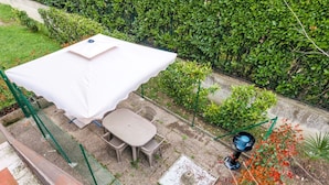 Un patio con mesa y sillas bajo un paraguas blanco, rodeado de follaje y un pequeño jardín.