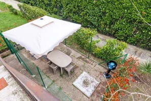 Un patio con mesa y sillas bajo un paraguas blanco, rodeado de follaje y un pequeño jardín.