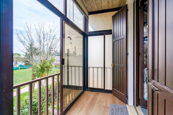 A balcony with glass doors, a wooden floor, and a view of a garden and buildings.