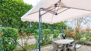 Un patio con mesa y sillas bajo una cubierta, rodeado de follaje y una cerca.