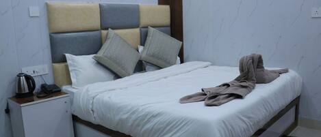 A neatly made bed with a white comforter and pillows, a bedside table with a kettle and phone, and a small cabinet.