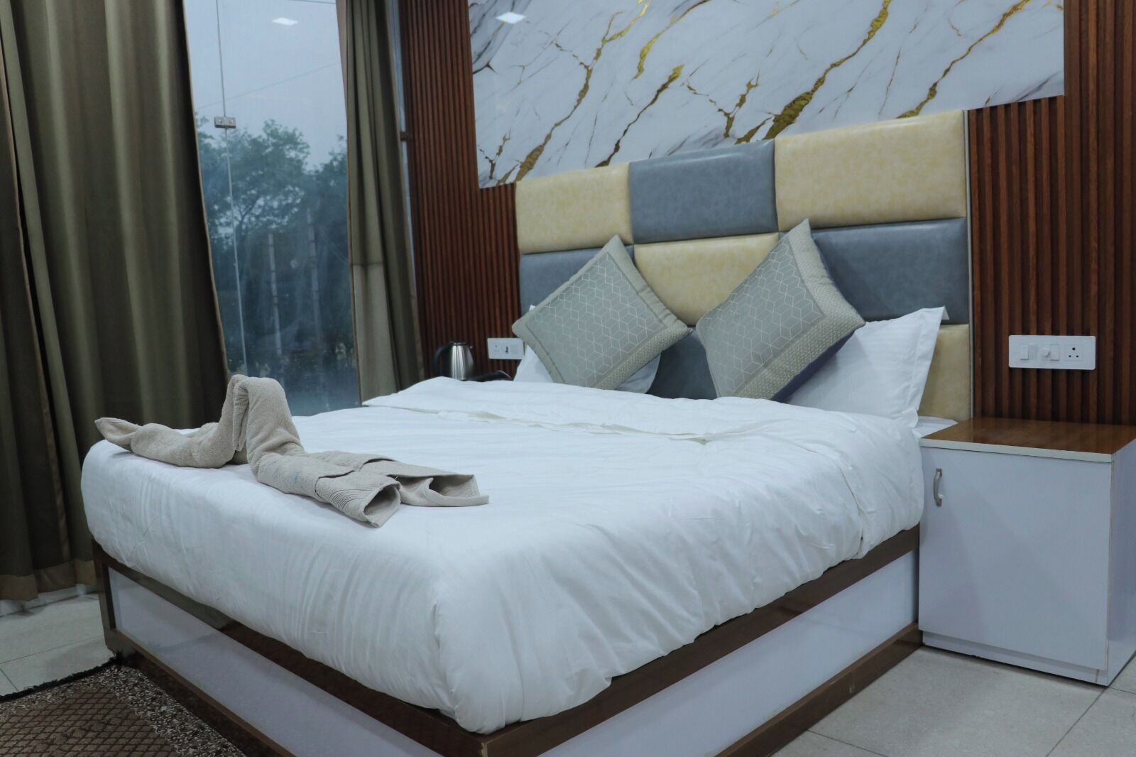 A neatly made bed with a white comforter and pillows, a wooden headboard with a marble pattern, a small bedside table, and a view of trees through a window.
