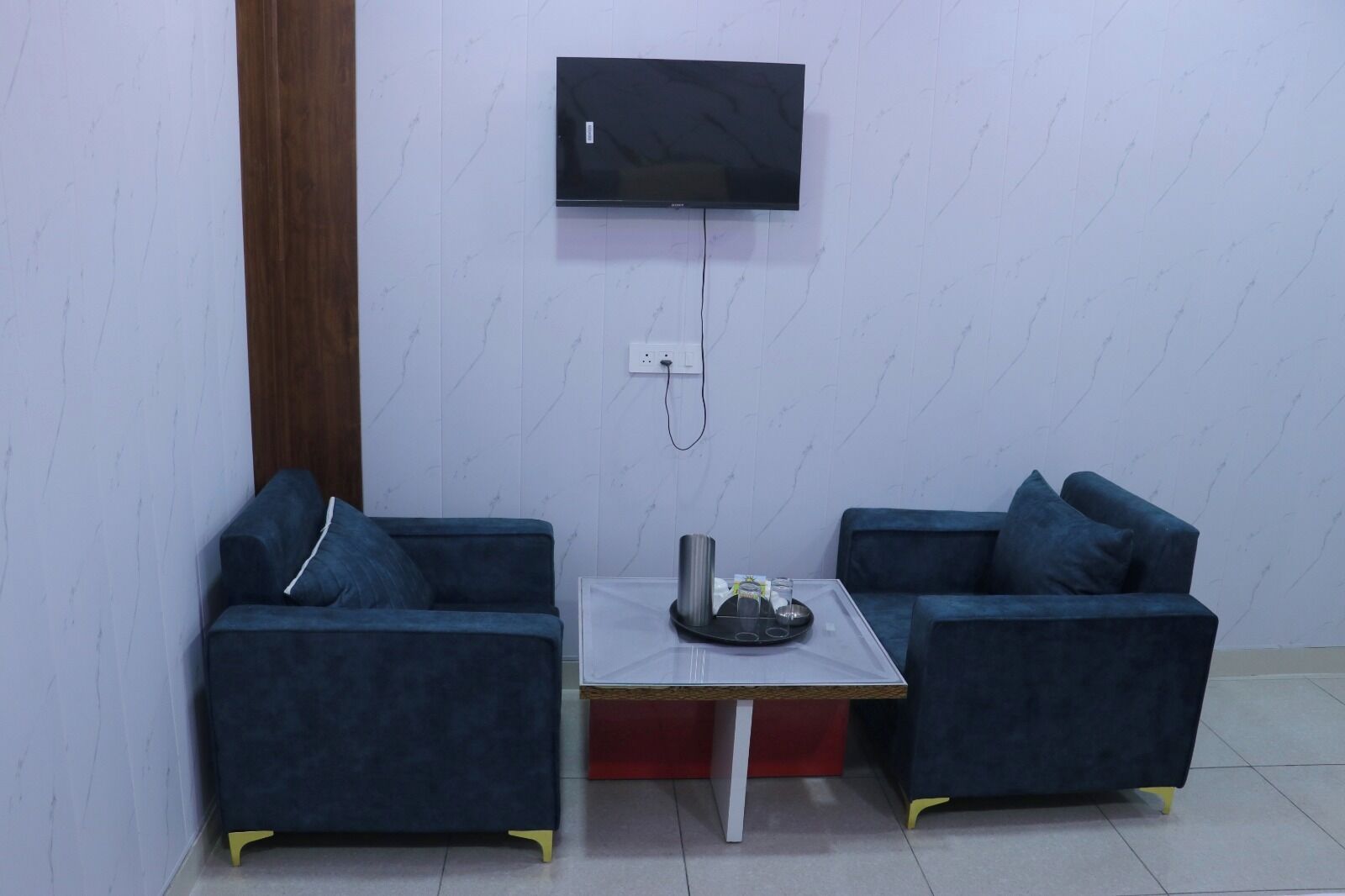 A room with two dark blue sofas, a small table with a tray, a television mounted on the wall, and a wooden door.