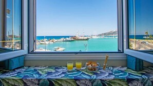 Blick aus dem Fenster auf eine Marina mit Booten, bewölkter Himmel und ein Frühstücksbuffet mit Saft und Gebäck auf einer blumigen Tischdecke.