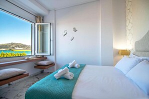 A hotel room with a bed, a window with a view of a coastal landscape, a bedside table with a lamp, and a wall-mounted decorative item.