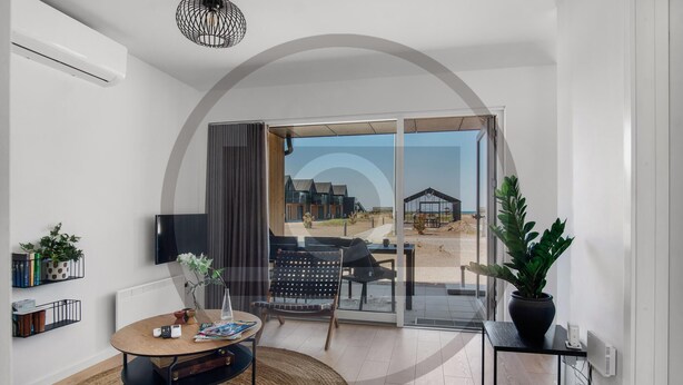 A modern living room with a large sliding glass door leading to a balcony, a round wooden table, a sofa, and a TV mounted on the wall. - Havnehusene, Lejl. 51 (Slagelse)