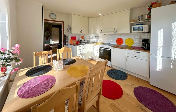 Une cuisine moderne avec des meubles blancs, un four à micro-ondes et un réfrigérateur. On y trouve une table à manger avec des chaises et des sets de table colorés.