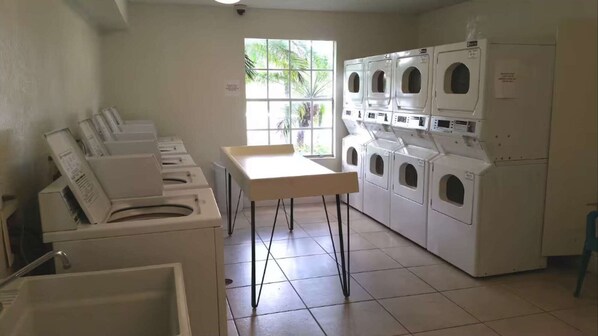Une buanderie équipée de plusieurs machines à laver et de plusieurs sèche-linge, avec une table et une fenêtre donnant sur des palmiers.