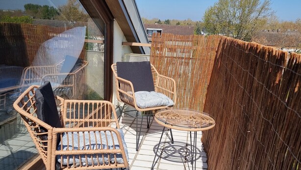 Een balkon met rieten meubels, een glazen tafel en uitzicht op de omgeving.