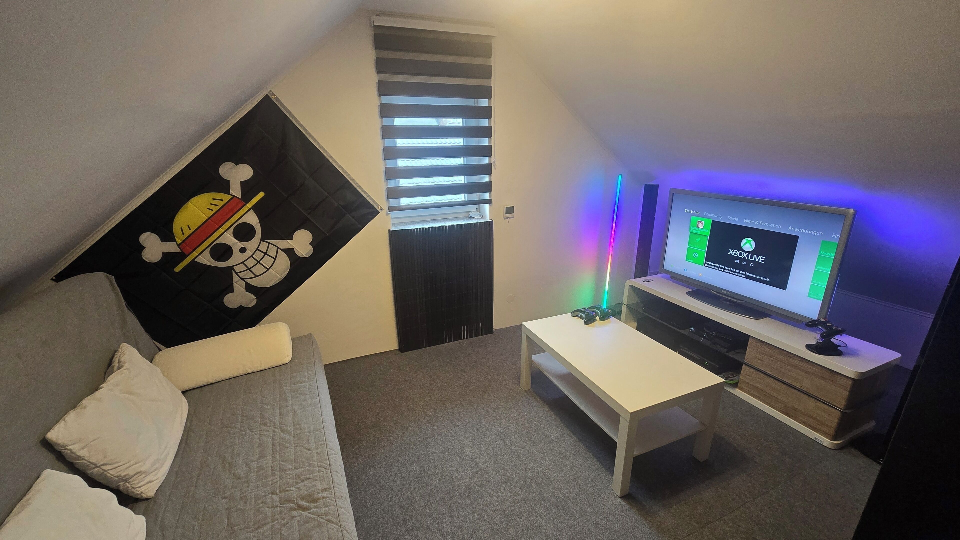 Ein kleiner, dreieckiger Raum mit einem Bett, einem Schreibtisch und einem Fernseher, der die Xbox Live-Oberfläche zeigt.