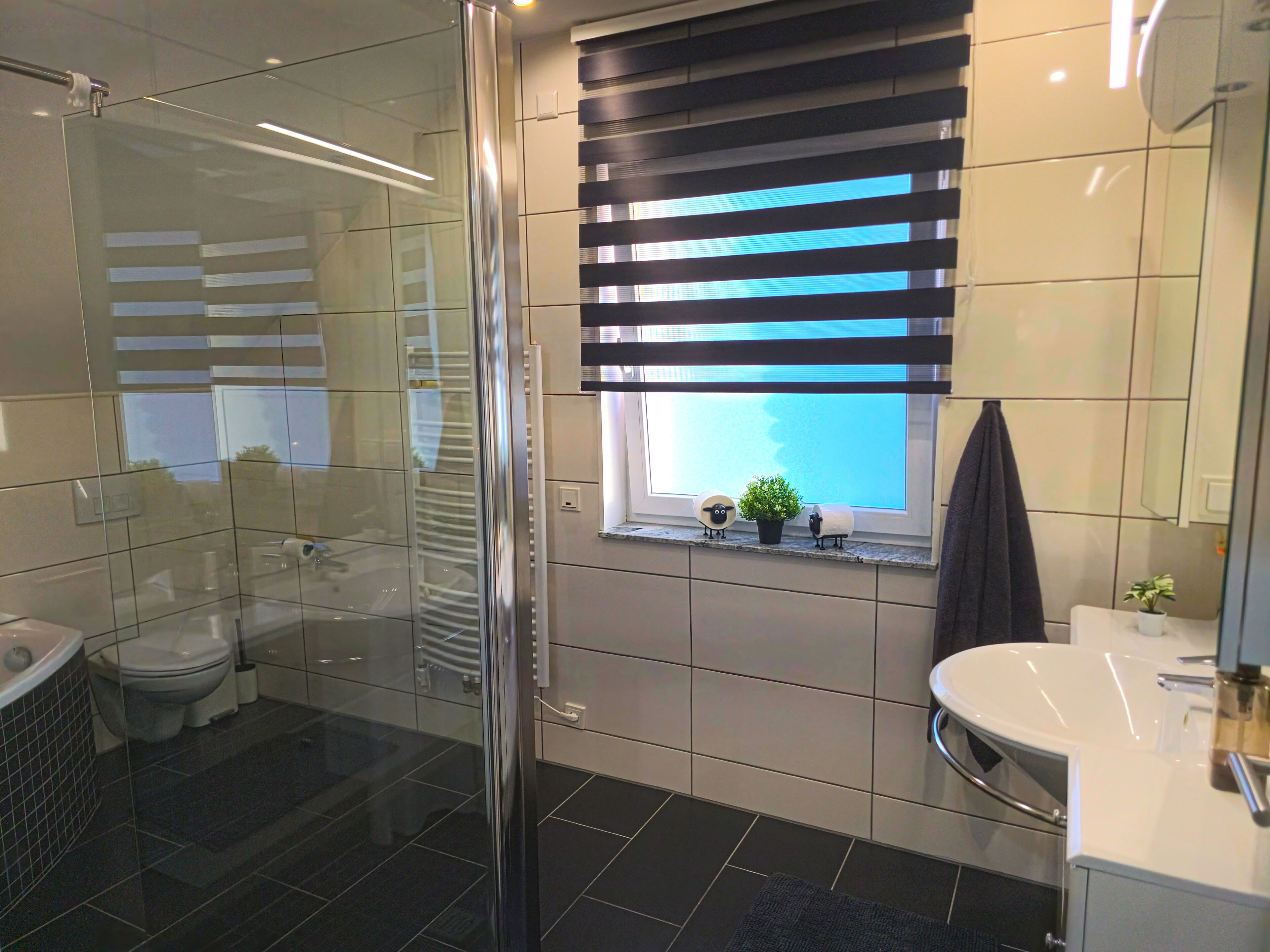 Ein modernes Badezimmer mit Duschkabine aus Glas, Toilette, Waschbecken und einem Fenster mit Jalousien.