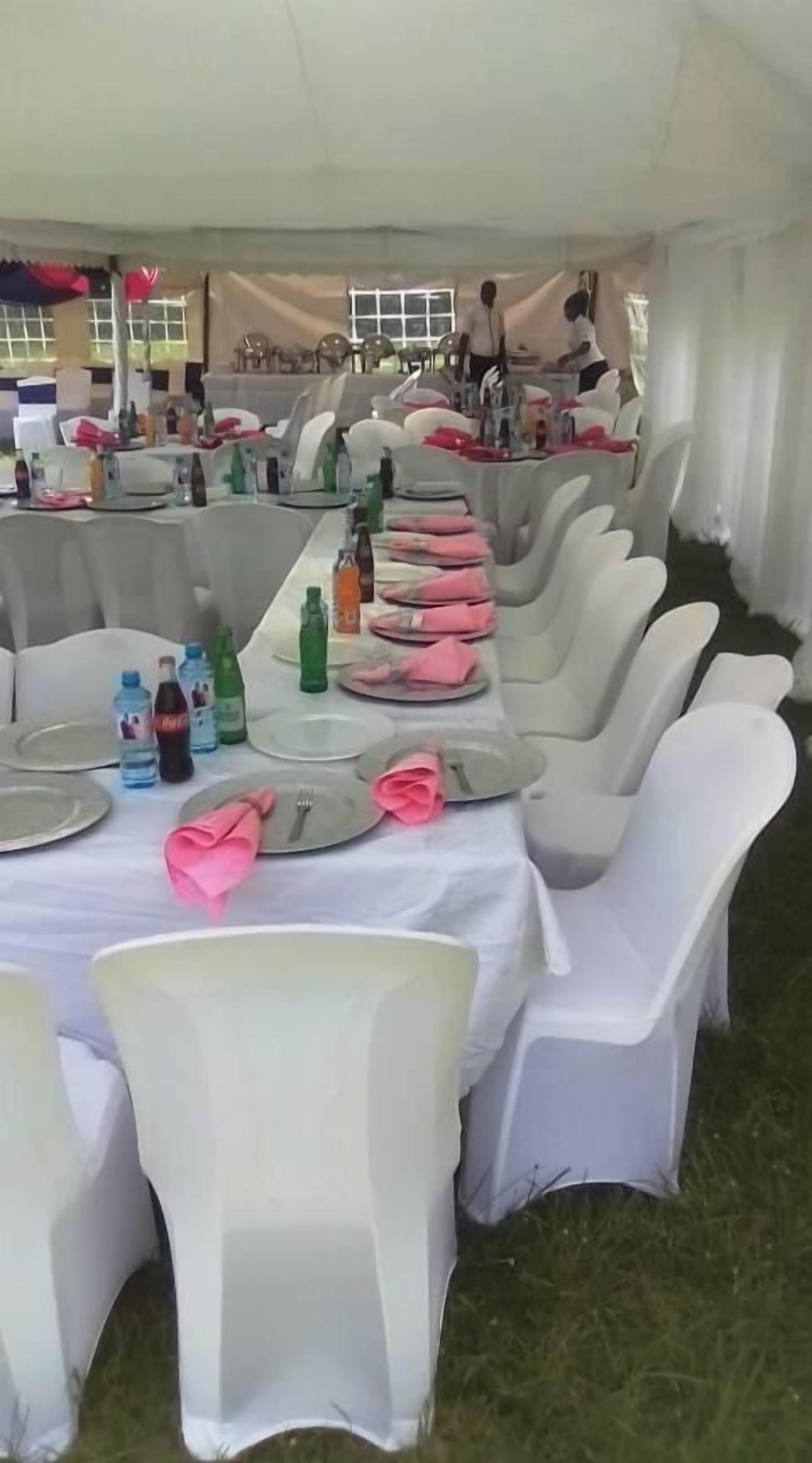 Un espacio al aire libre con carpas, mesas dispuestas para una comida, manteles blancos y sillas, y diversas bebidas sobre las mesas.