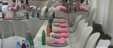 Un espacio al aire libre con carpas, mesas dispuestas para una comida, manteles blancos y sillas, y diversas bebidas sobre las mesas.