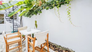 Een klein buitenterrein met een witte tafel en houten stoelen, omgeven door weelderige begroeiing en een witte muur.