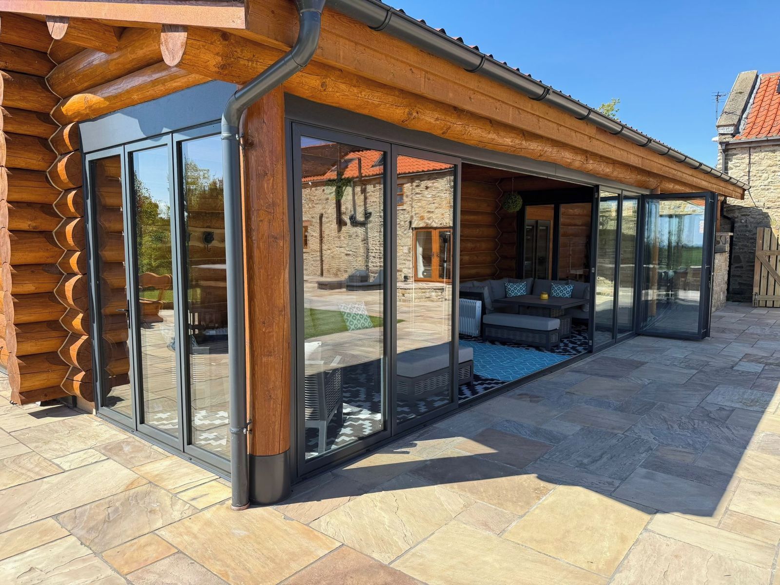 A modern log cabin with large sliding glass doors, a stone exterior, and a patio area with outdoor furniture.