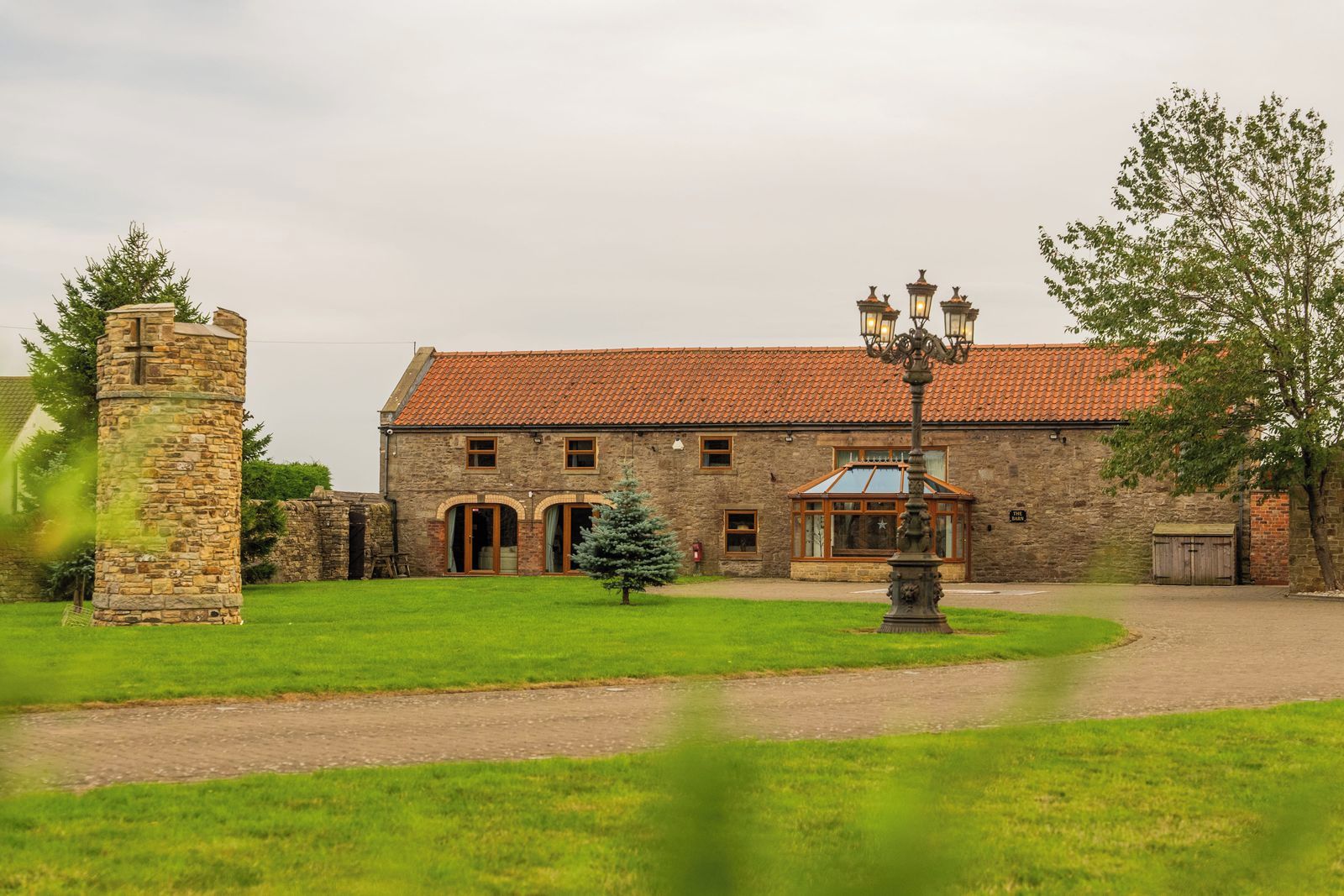 A stone building with a red-tiled roof, a central entrance with arched doorways, and a small tree in the foreground.