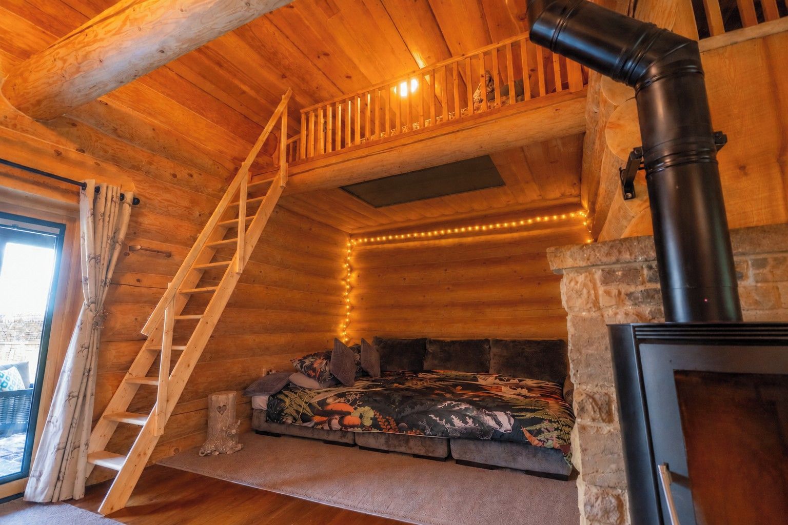 A cozy cabin interior with a wooden staircase, a fireplace, and a window overlooking a snowy landscape.