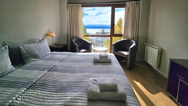 Habitación de hotel con una cama grande, dos sillas, una mesita, una lámpara y vistas al exterior.