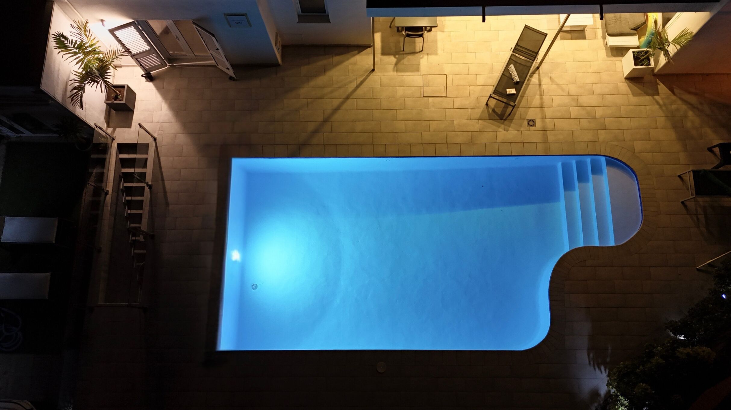 A top-down view of a swimming pool with a blue tint, surrounded by a brick wall and a staircase.