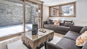 Ein modernes Wohnzimmer mit einer Steinwand, einem Glastisch, einem grauen Sofa und einem Fenster mit Blick auf eine verschneite Landschaft.