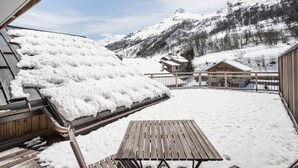 Eine Holzterrasse mit Tisch und Stühlen, im Hintergrund verschneite Berge und ein Gebäude mit einem geneigten Dach.
