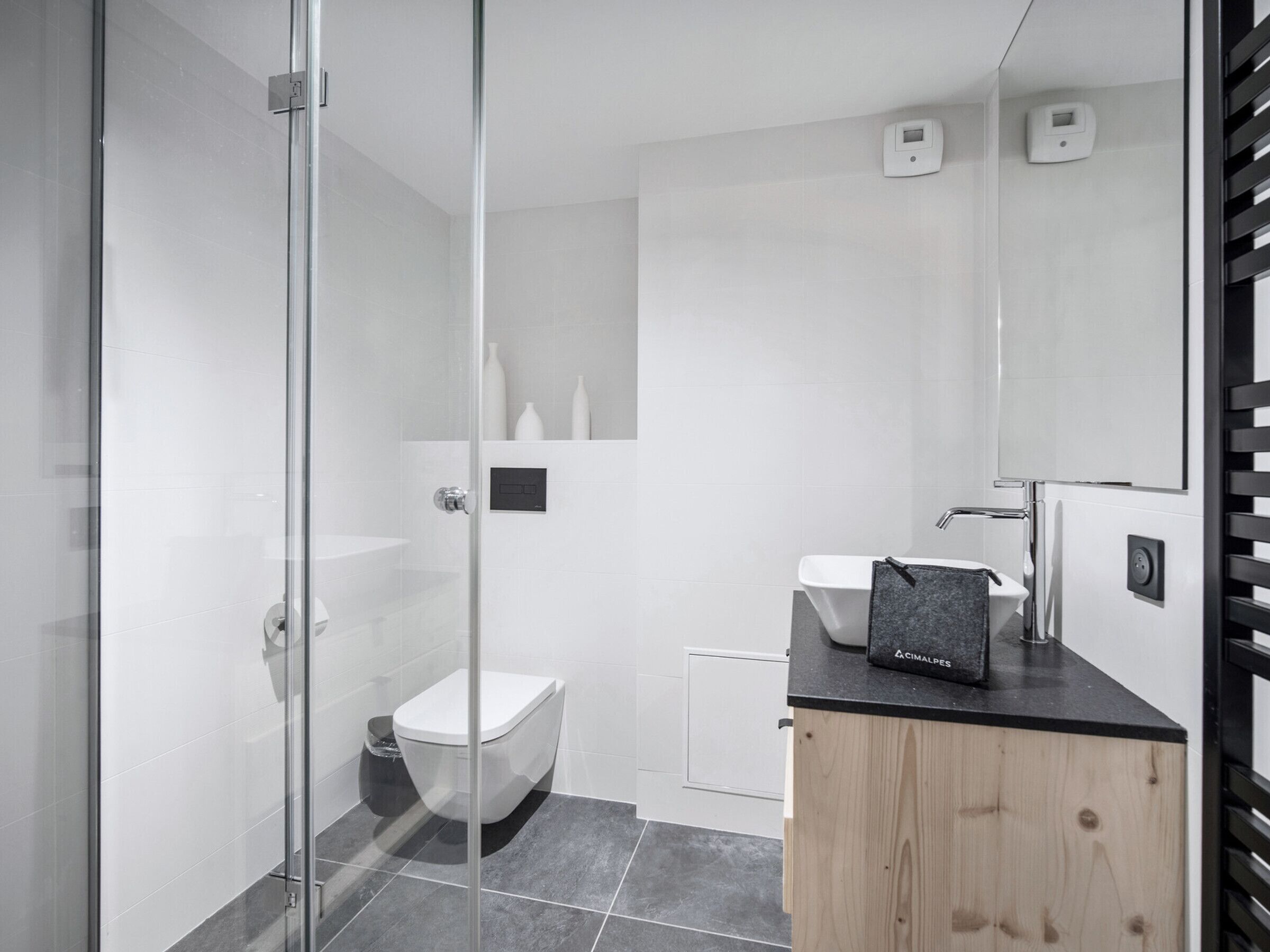 Et moderne badeværelse med glasbrusekabine, hvidt toilet, vask med sort bordplade og et træskab.