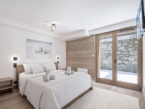 A modern bedroom with a large bed, a wooden wall panel, a stone wall, and a sliding glass door leading to a snowy outdoor area. - New apartment near slopes - 3 Valleys, parking included (Saint-Martin-de-Belleville)