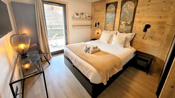 A bedroom with a bed, a bedside table with a lamp, a wooden headboard, and a view of the outside.